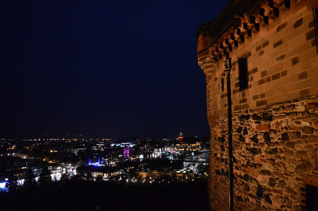 Mur du château au 1er plan, éclairé par la lumière, avant la ville et ses lumières au loin en contrebas dans la nuit