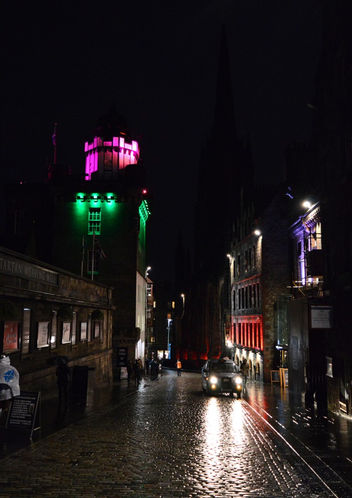 Rue pavée de nuit, avec éclairage multicolore des bâtiments, et un taxi stationnant 