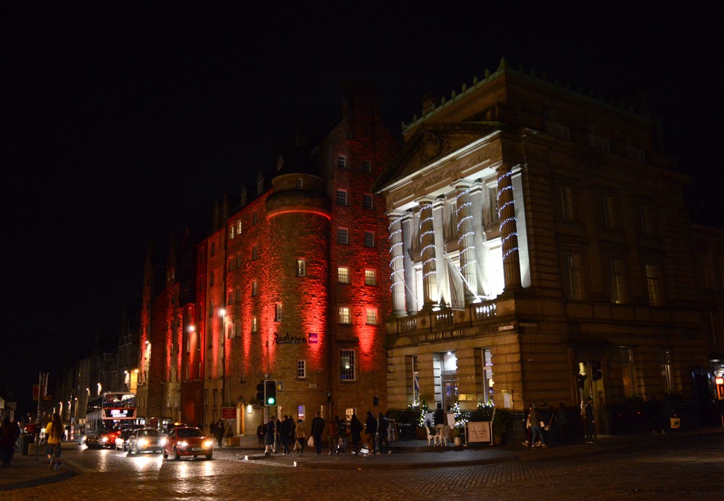 Décorations de noël sur les façades : bâtiment éclairé en rouge, avec des voitures passent devant, sur la route pavée