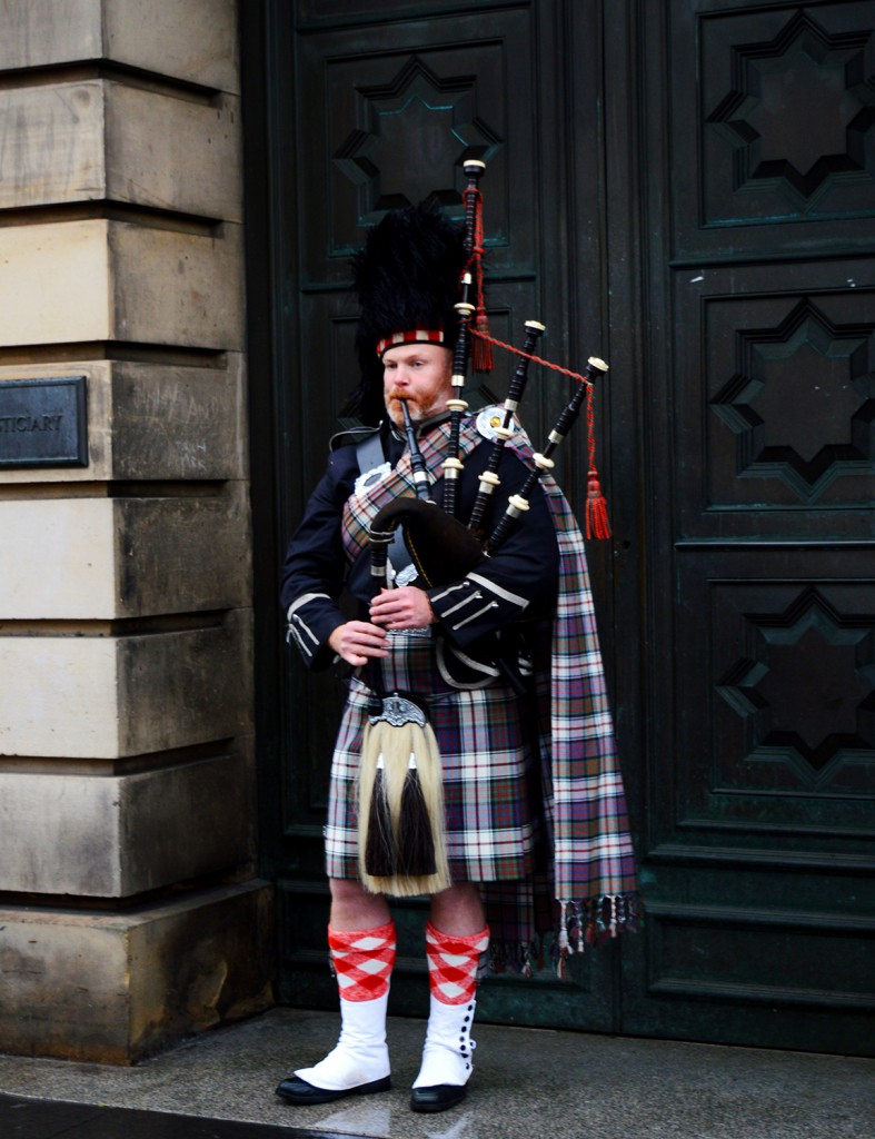 Personne en kilt et tartan à carreaux, jouant de la cornemuse au pied d'une grande porte en bois, dans la rue