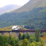 Un train à vapeur noir, vu dans Harry Potter, traverse un viaduc de pierre aux multiples arches, entouré d'arbres et de collines verdoyantes en Écosse, sous un ciel nuageux.
