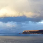 Une falaise côtière s'avance dans une mer calme et bleue sous d'épais nuages lors du coucher de soleil.