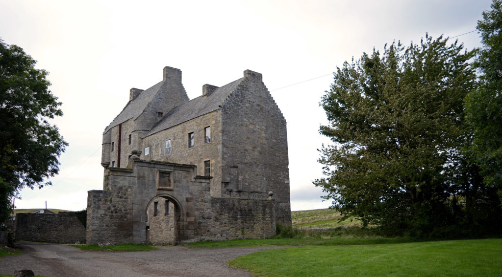 La maison de lallybroch, vue dans son ensemble, entre 2 grands arbres, bordée par une pelouse verte