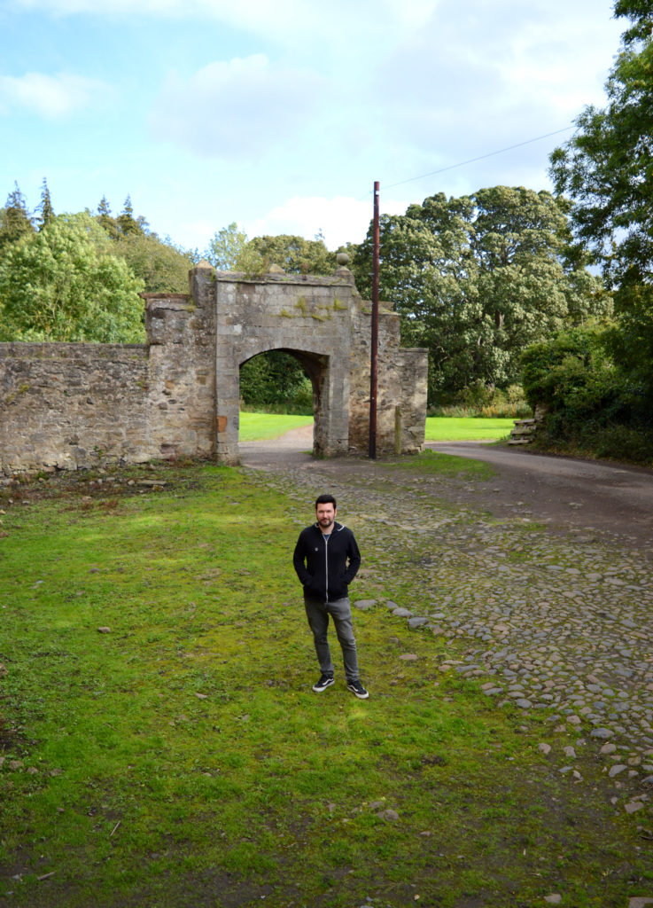 Manu debout sur l'herbe verte, devant l'entrée de Lallybroch