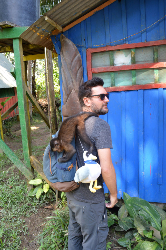 Manu debout, avec un singe capucin dans le dos sur son sac à dos, en train de fouiller les poches du sac