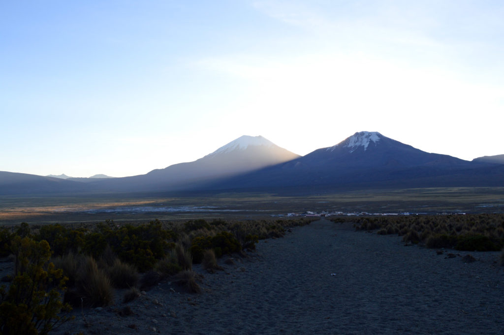 Chemin de cailloux dans la plaine, menant vers le village. Les volcans Parinacota et Pomerape sont en arrière plan, avec le Parinacota éclairé de la lumière du coucher de soleil. Le Pomerape est dans l'ombre