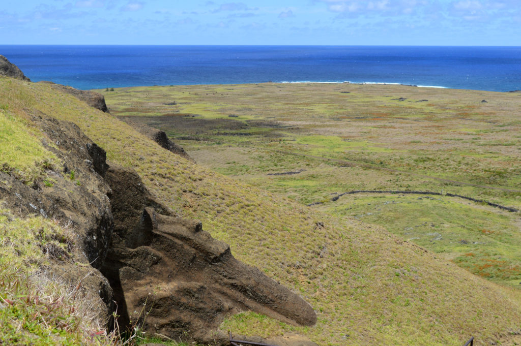 Vue depuis la montagne sur un moaï creusé dans la falaise, avec l'océan en arrière plan