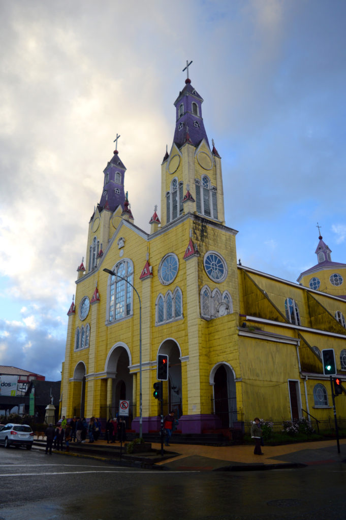 Église en bois, peinte en jaune, et avec des clochers de couleur violette, devant un ciel menaçant mais partiellement bleu