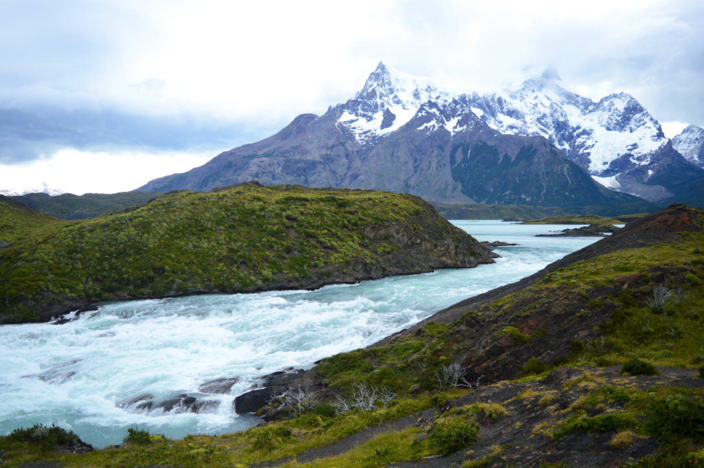 Eau agitée, de couleur bleue glace, d'une rivière formant une petite cascade sur des rochers. Les berges sont vertes, et on peut apercevoir des sommets enneigés au loin