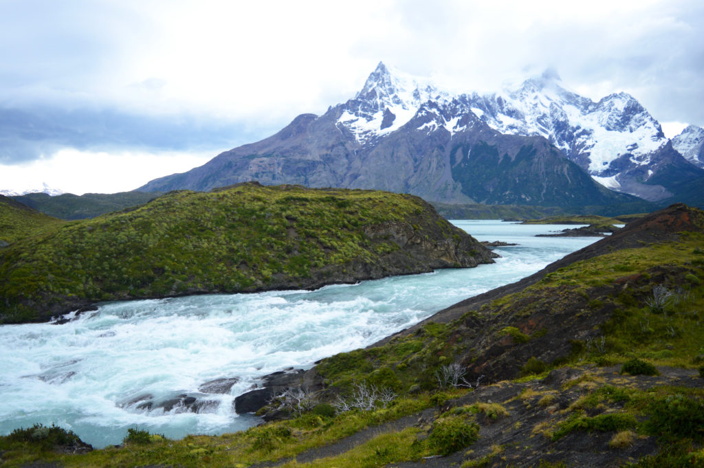 Eau agitée, de couleur bleue glace, d'une rivière formant une petite cascade sur des rochers. Les berges sont vertes, et on peut apercevoir des sommets enneigés au loin