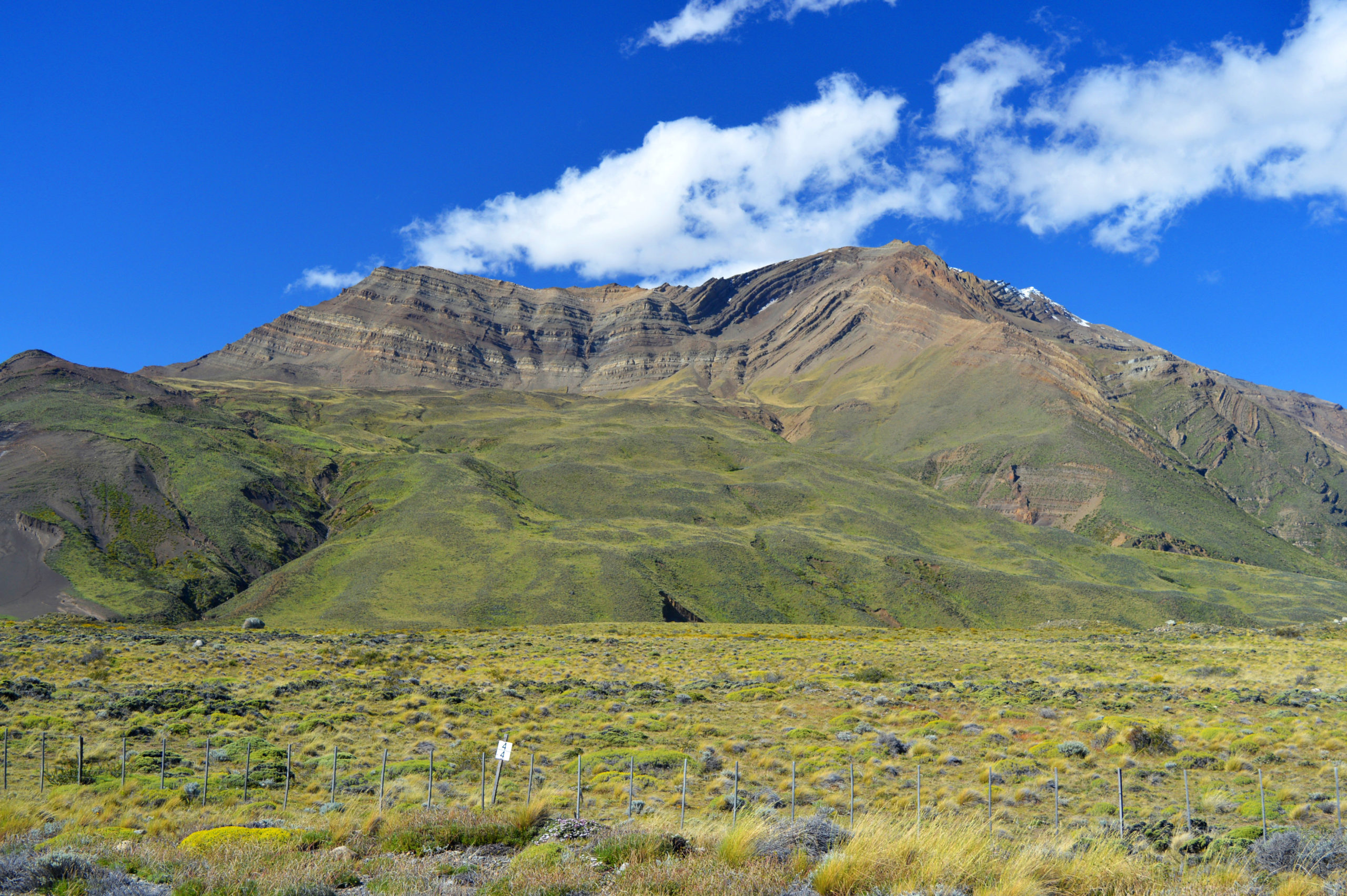 Une grande montagne rocheuse s'élève au-dessus d'une plaine d'herbe sous un ciel bleu vif avec des nuages blancs épars