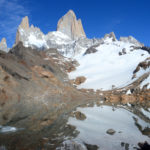 Les pics montagneux enneigés s'élèvent au-dessus d'un terrain rocheux et d'un lac clair, qui reflète les montagnes et le ciel bleu, vue mythique lors de la randonnée à El Chalten