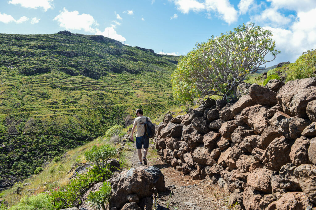 Manu marchant sur un chemin à flanc de colline, bordé par de grosses pierres volcaniques. Au loin une colline verte à stries