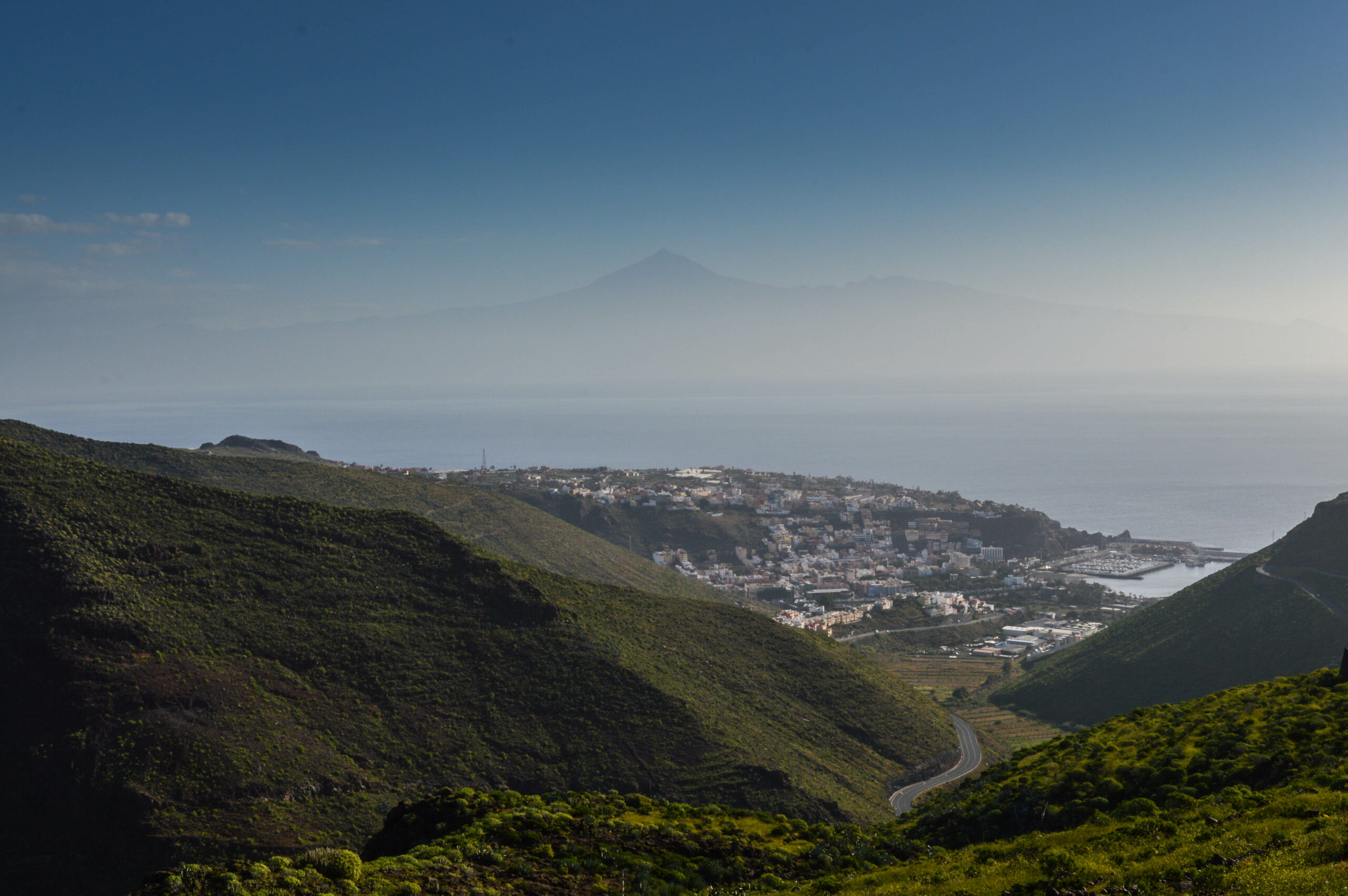 Vue en hauteur de San Sebastian de la gomera : collines verdoyantes au premier plan, ville au second plan, et le contour au loin, derrière la mer, du volcan Teide