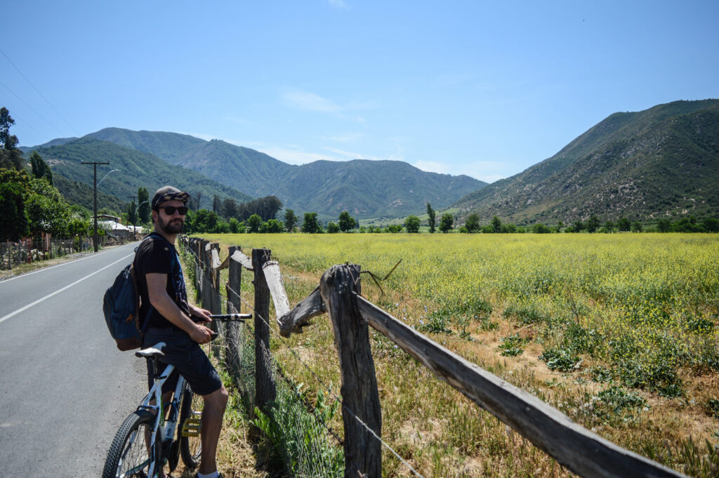 Manu à vélo, en bord de route, dans la vallée, avec les montagnes au fond