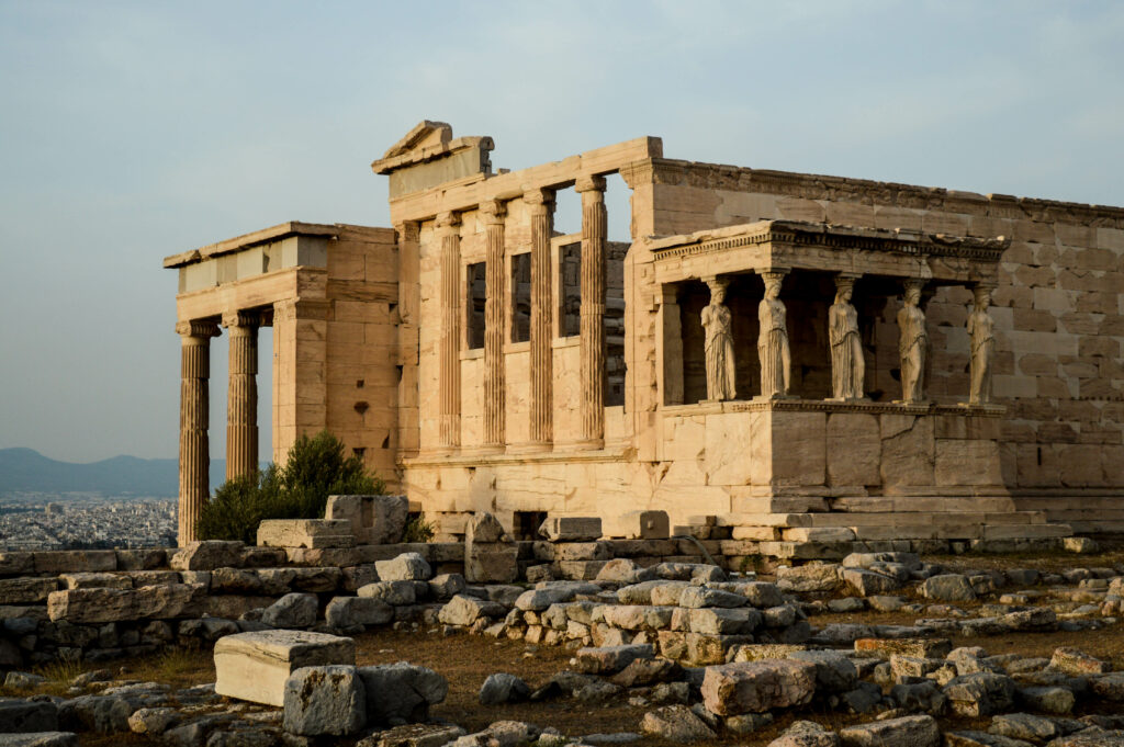 Vue lointaine sur le temple d'Érechthéion, avec de hautes colonnes et six statues de femmes soutenant une partie du toit, entourée de ruines éparses et d'un sol rocailleux sous un ciel nuageux.
