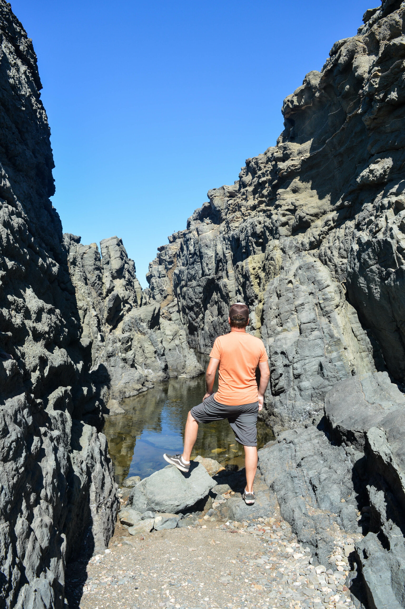 Une personne vêtue d'une chemise pêche et d'un short gris se tient sur un rocher, face à un canyon étroit aux hautes falaises escarpées et à une mare d'eau peu profonde