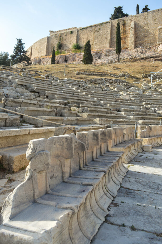 Des rangées d'anciens sièges en pierre forment l'amphithéâtre du théâtre de Dionysos sur une colline, avec un mur de pierre et quelques grands arbres au-dessus. La scène éclairée par le soleil met en valeur la texture altérée de ces ruines historiques.