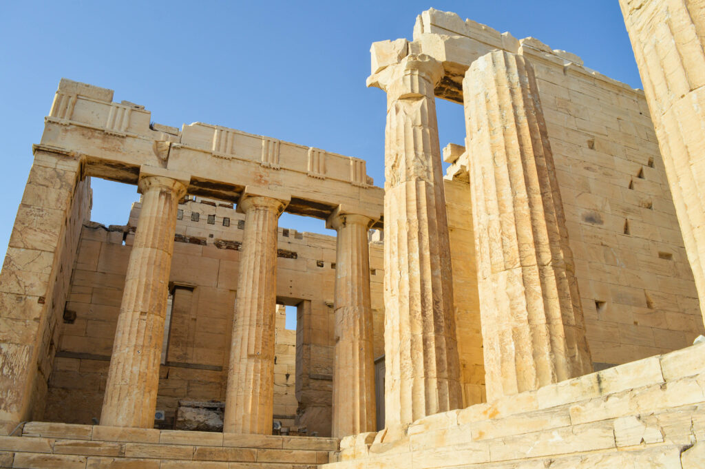 Vue rapprochée de ruines grecques antiques à l'acropole, avec de hautes colonnes de pierre cannelées et des murs usés par les intempéries, sous un ciel bleu clair. La structure semble partiellement intacte et témoigne d'un style architectural grec classique.