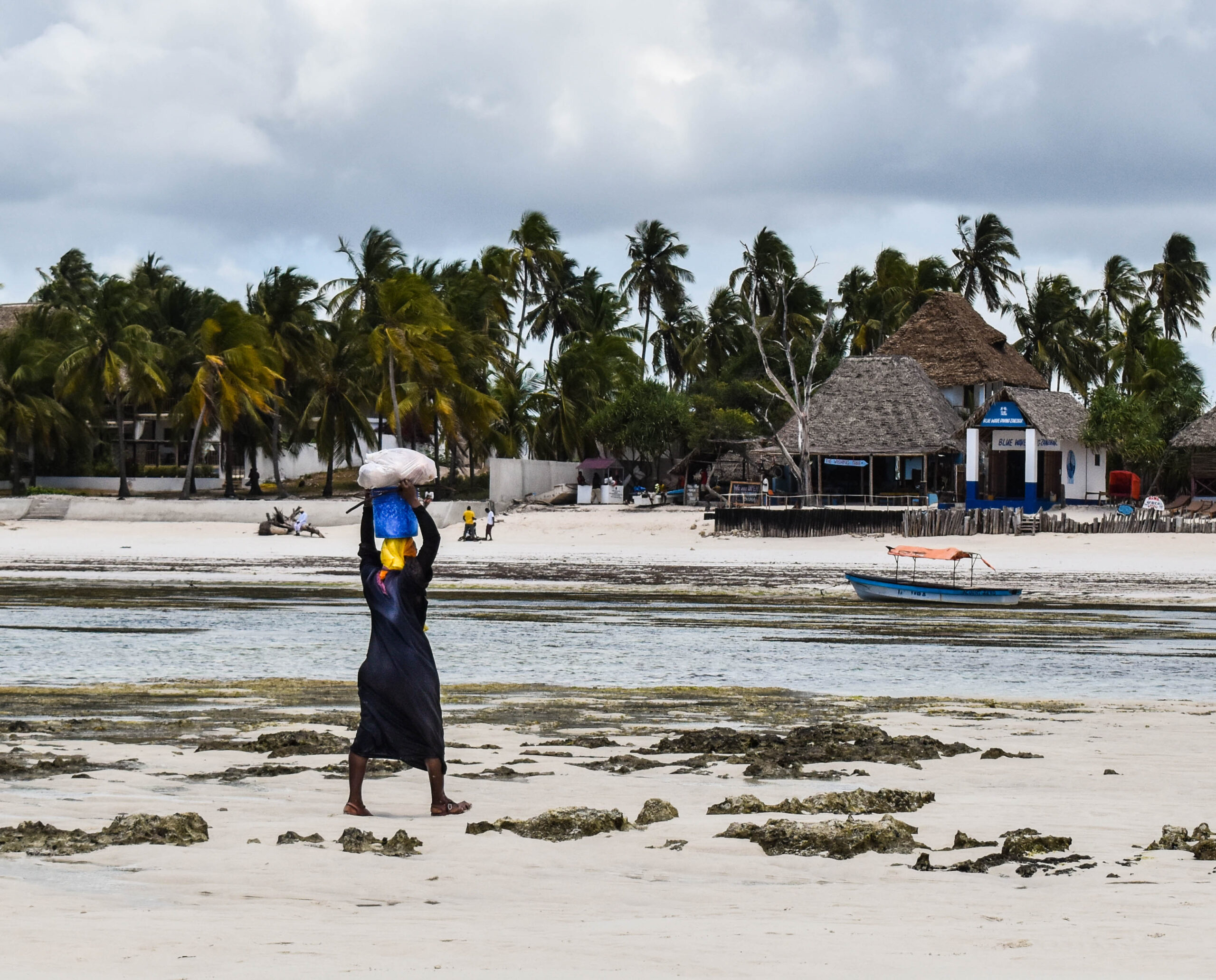 Femme de dos, qui porte un sac sur sa tête, sur la plage à Zanzibar. Au loin des palmiers et quelques paillotes