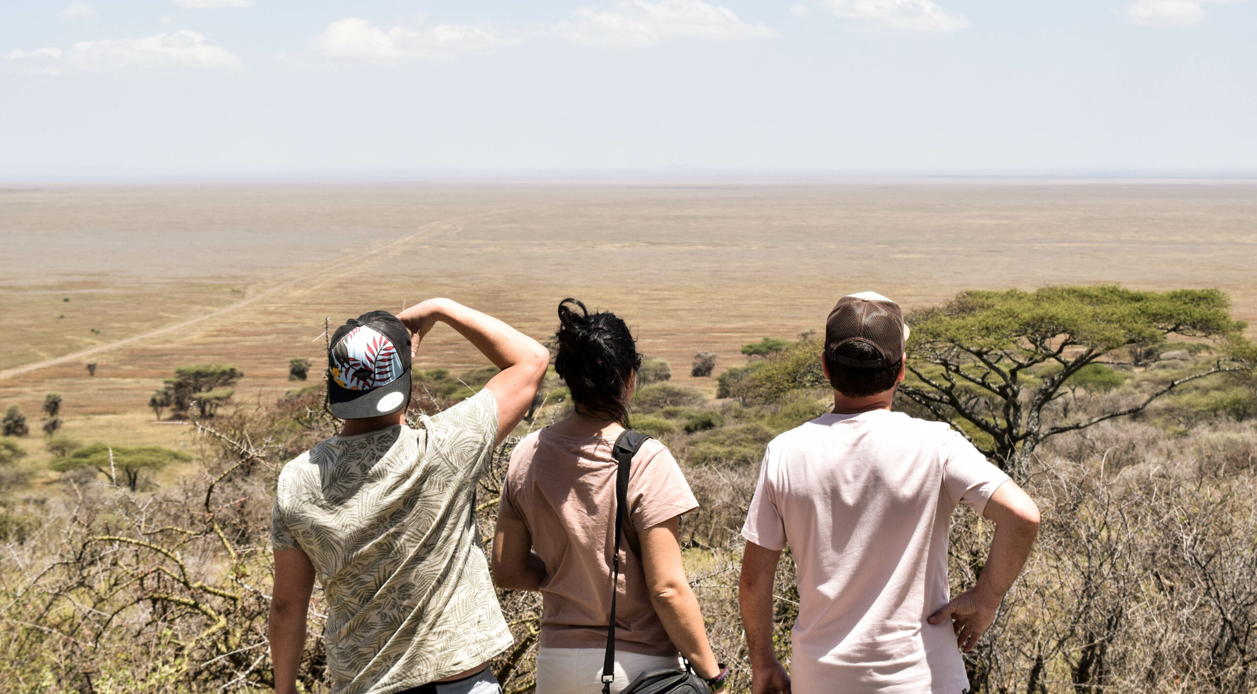 3 personnes de dos, en hauteur, observant à perte de vue la plaine du Serengeti, avec quelques acacias au 1er plan
