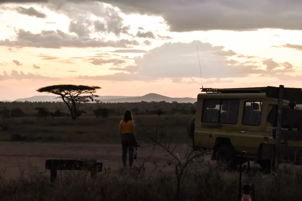 Audrey devant le coucher de soleil sur la savane à côté d'une jeep