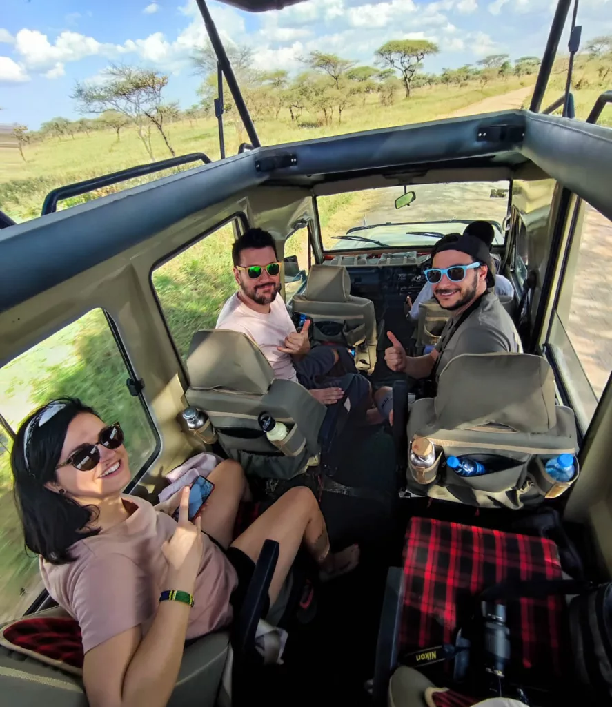 Amandine Manu et Thomas dans le 4x4, souriants, avec le paysage de la savane visible grâce au toit ouvrant