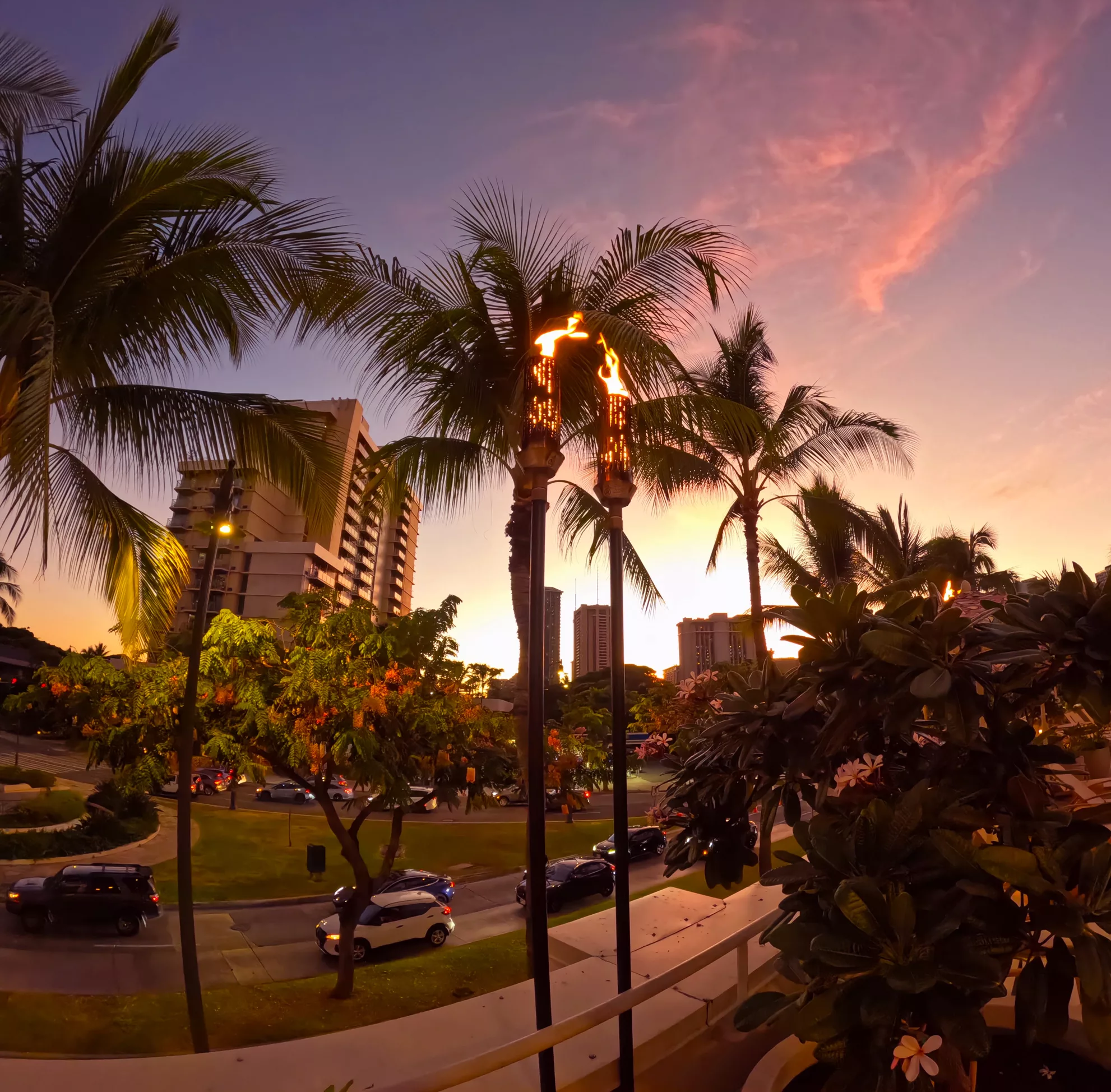 Une rue à Honolulu Hawaï, bordée de palmiers et de plantes à fleurs au coucher du soleil. Les voitures passent devant de grands immeubles tandis que deux torches tiki flamboyantes brillent au premier plan.