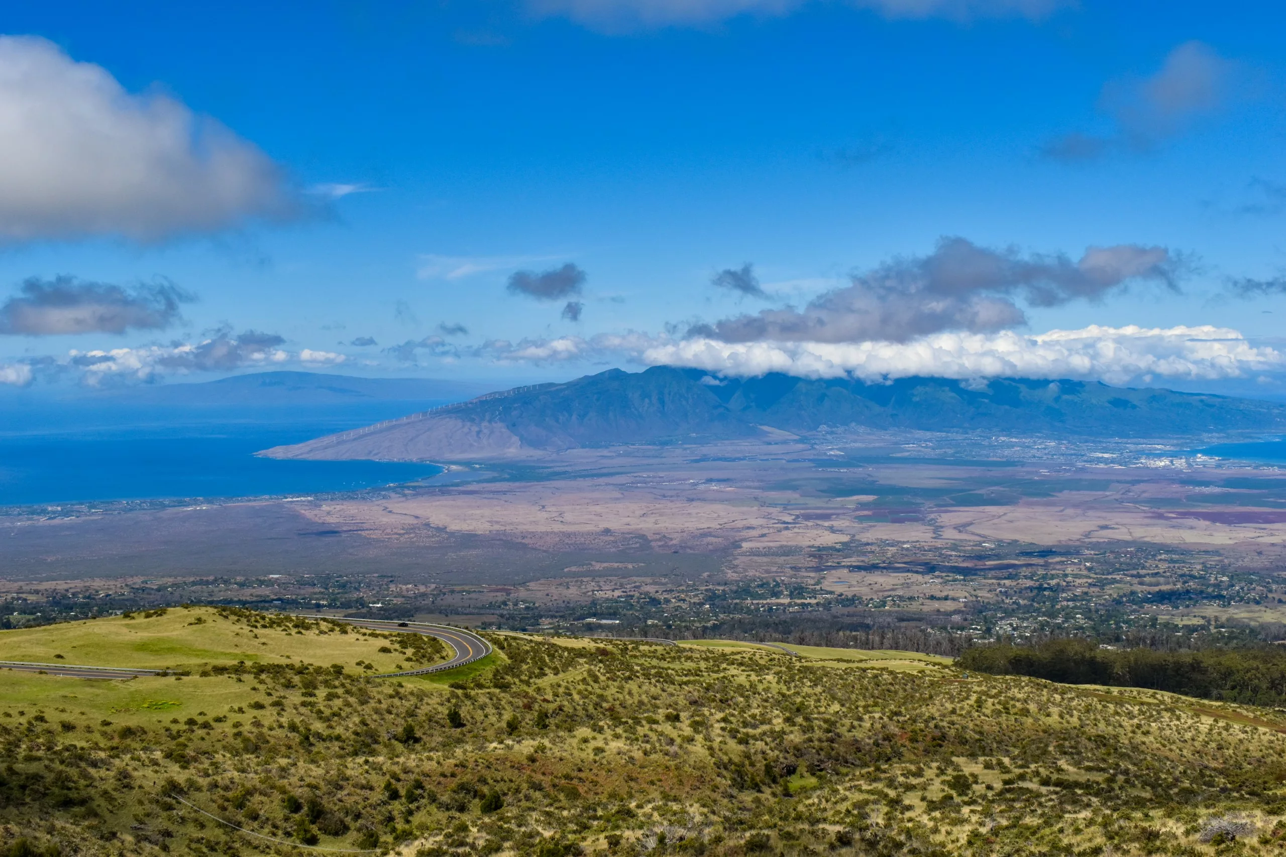 Une vue panoramique sur Maui et son paysage verdoyant avec une route sinueuse, des plaines côtières et des montagnes lointaines sous un ciel bleu