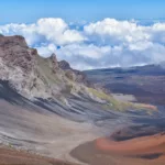 Un large cratère volcanique à Haleakala avec des pentes rocheuses multicolores et des taches de végétation verte sous un ciel bleu vif, avec des nuages remplissant l'arrière-plan au-dessus du bord du cratère.