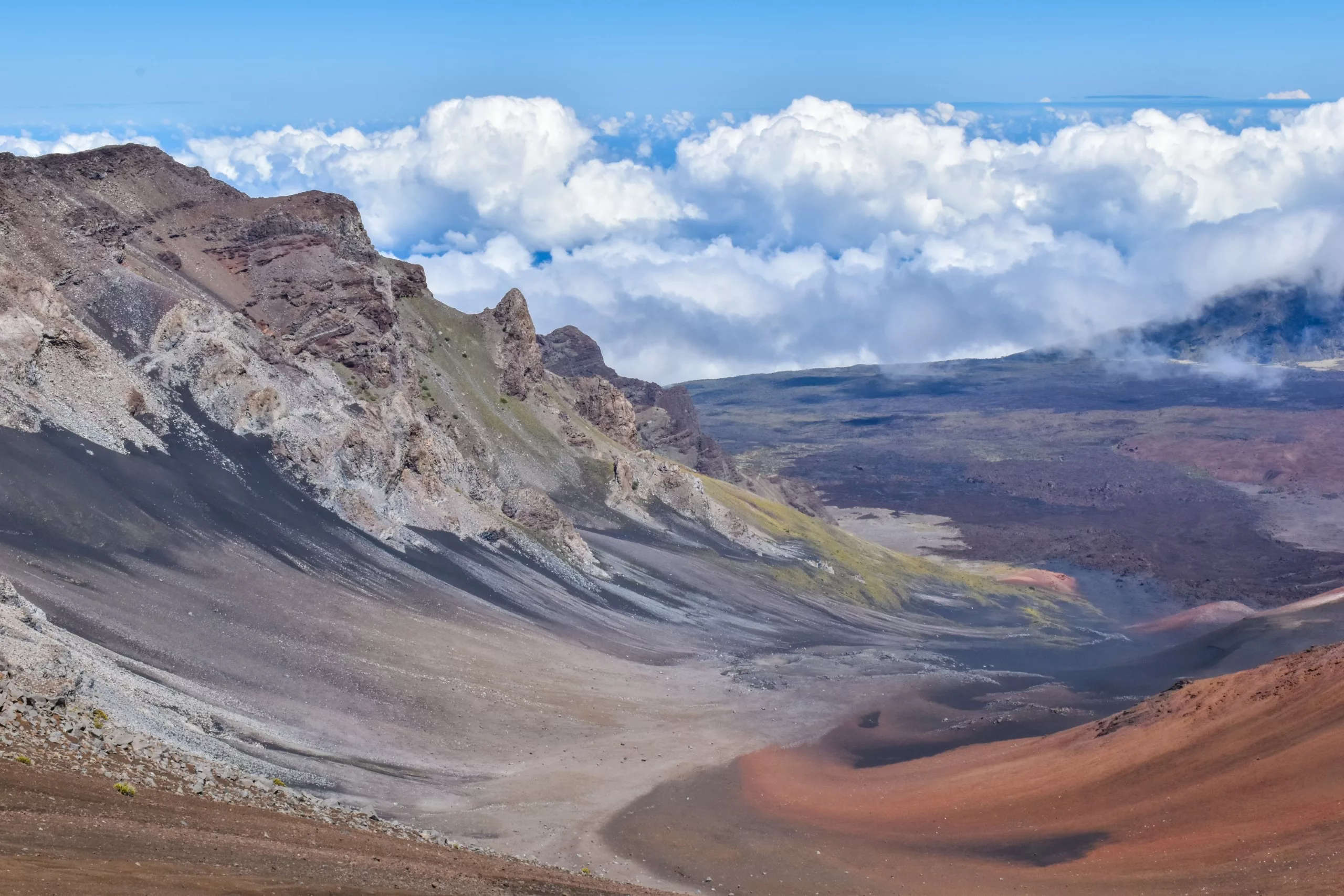 Un large cratère volcanique à Haleakala avec des pentes rocheuses multicolores et des taches de végétation verte sous un ciel bleu vif, avec des nuages remplissant l'arrière-plan au-dessus du bord du cratère.
