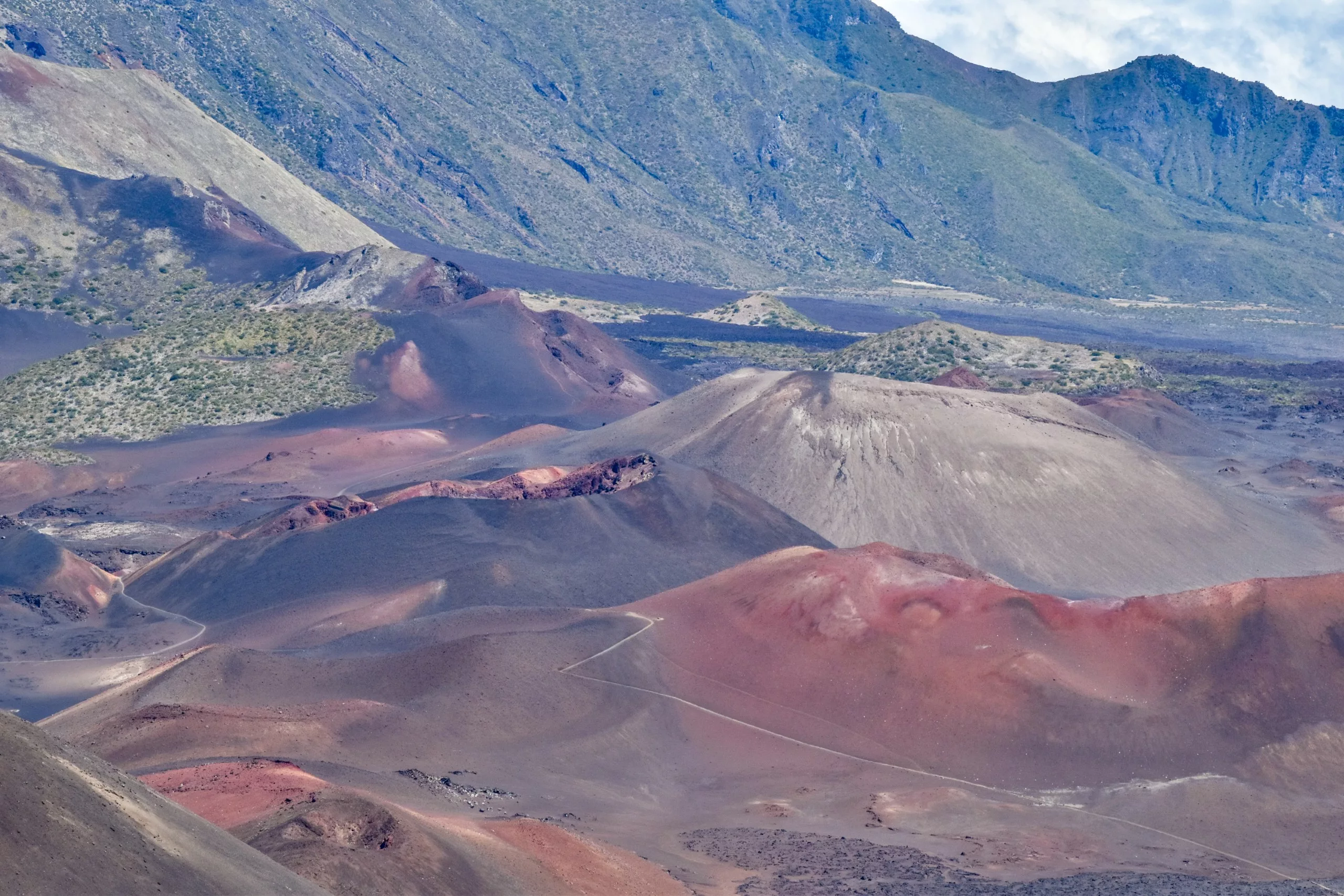 Des cônes de scories volcaniques colorés et un terrain rocheux remplissent ce paysage aride - l'un des sites incontournables à faire à Maui - sur fond de montagnes escarpées et accidentées sous un ciel partiellement nuageux.
