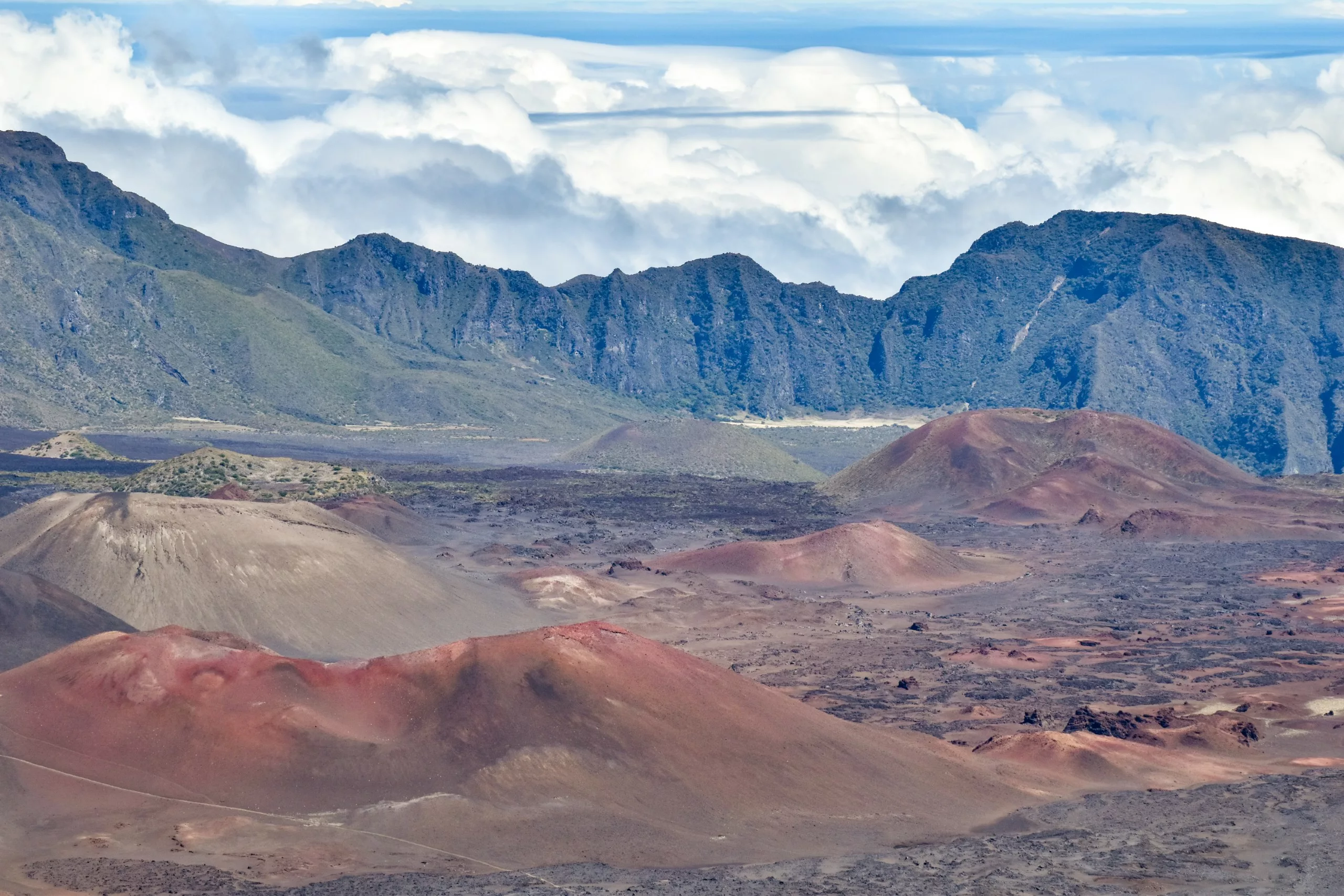 Des cônes de cendres volcaniques colorés et un terrain rocheux remplissent le premier plan -un incontournable si vous vous demandez ce qu'il faut faire à Maui - avec des falaises sombres et abruptes et une couche de nuages blancs sous un ciel bleu clair.