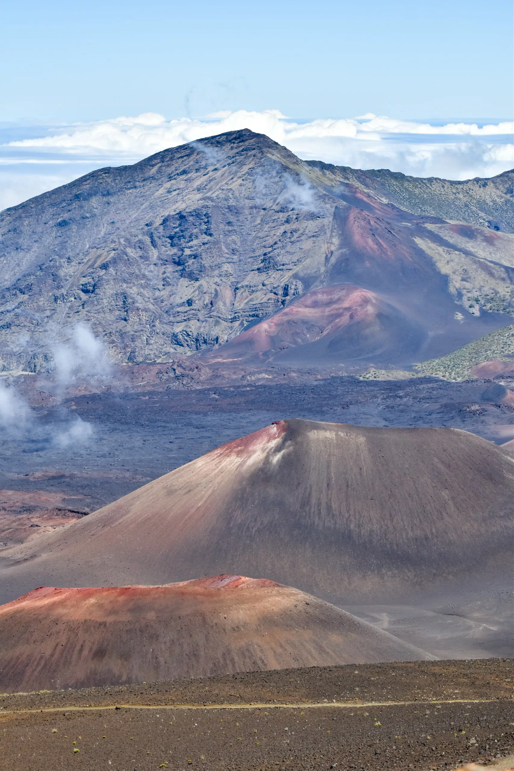 Un paysage volcanique aride avec plusieurs cônes de scories au premier plan et la grande montagne rocheuse de Haleakala en arrière-plan sous un ciel clair avec quelques nuages épars.