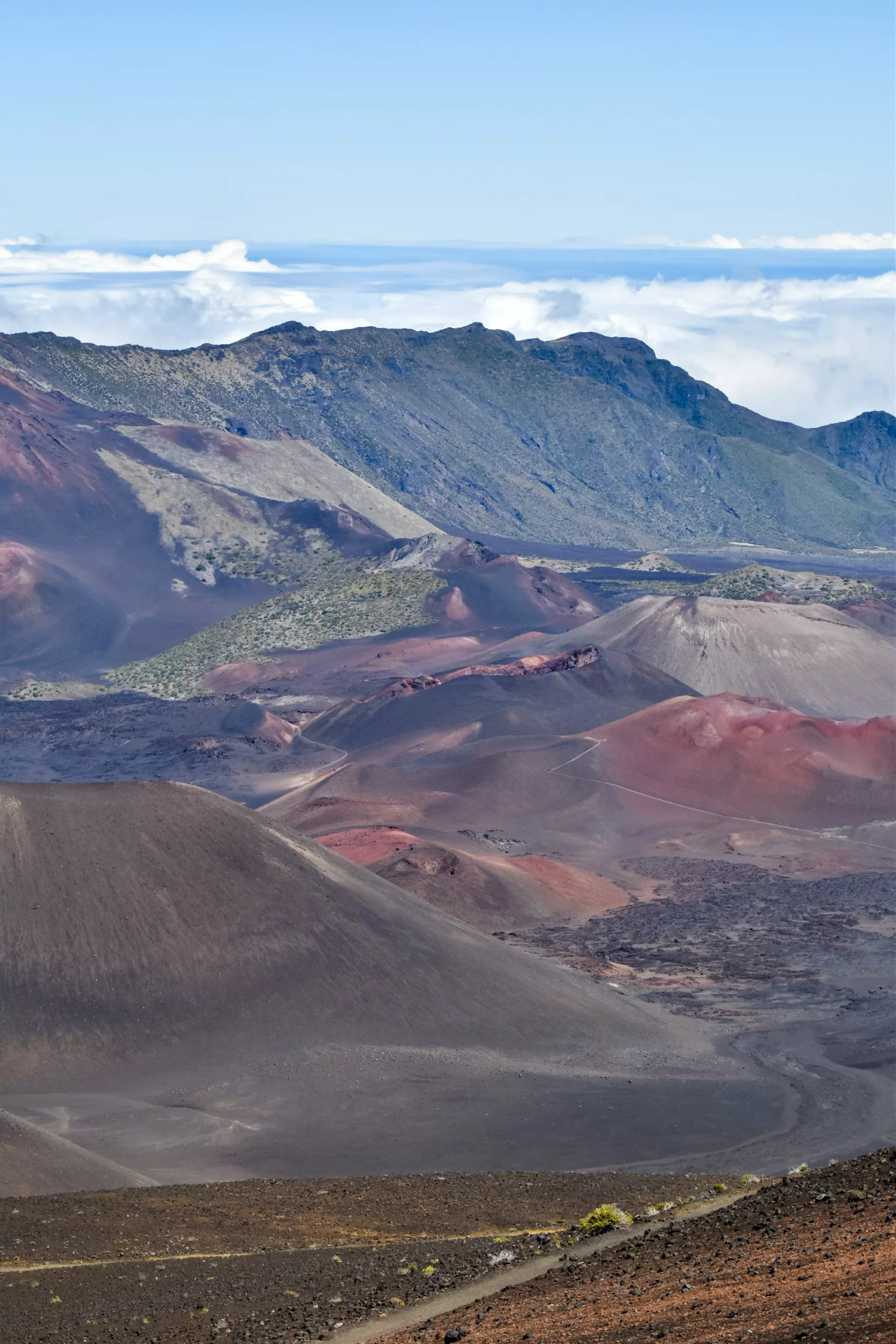 Une vue de paysage d'un cratère volcanique avec des pentes rougeâtres et brun foncé, un terrain rocheux, une végétation dispersée et des crêtes de montagnes lointaines sous un ciel bleu - une vue emblématique lors de l'exploration de ce que faire à Maui.