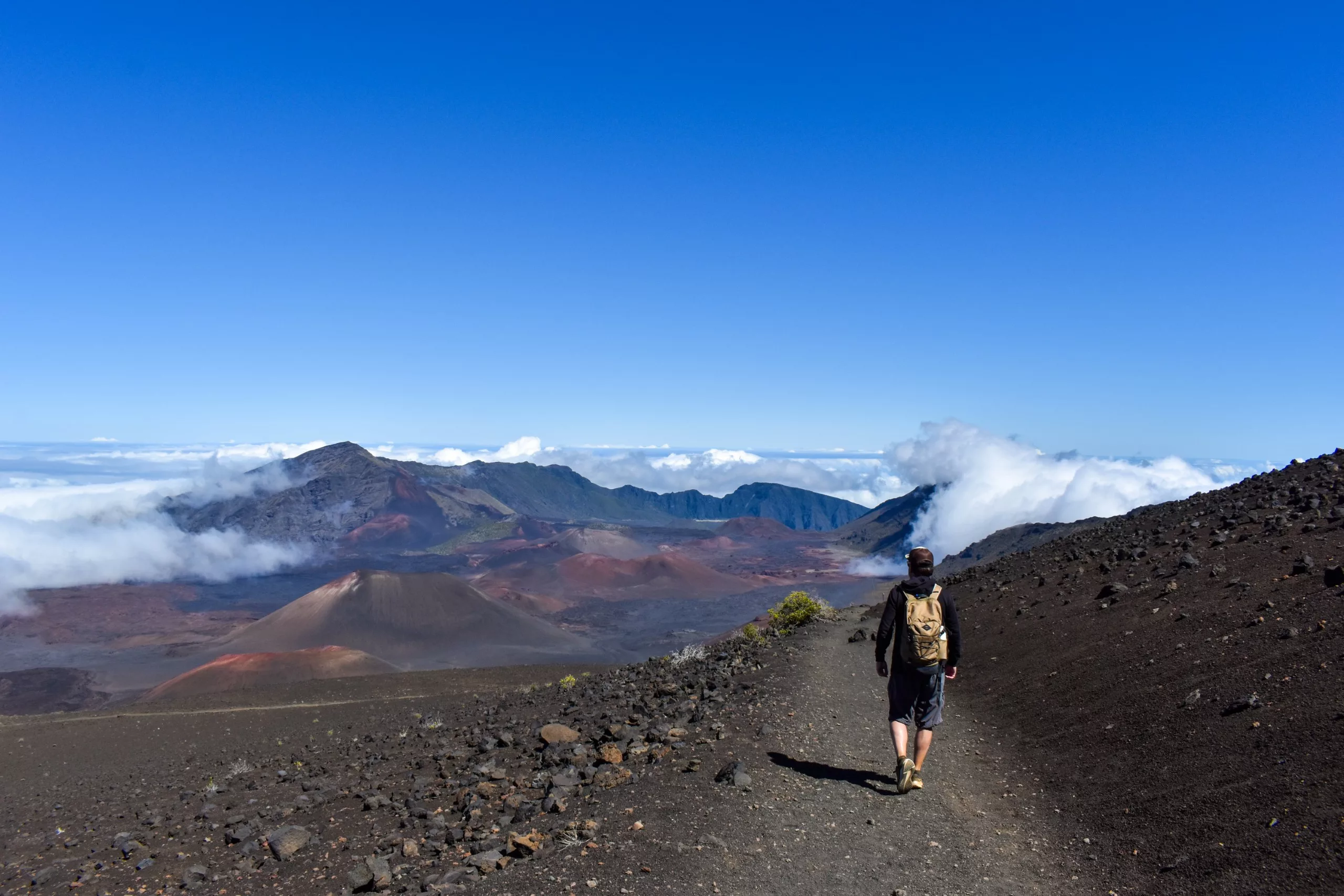Une personne avec un sac à dos marche le long d'un sentier dans le paysage volcanique de Haleakala, avec des montagnes, des nuages et un ciel bleu clair en arrière-plan.