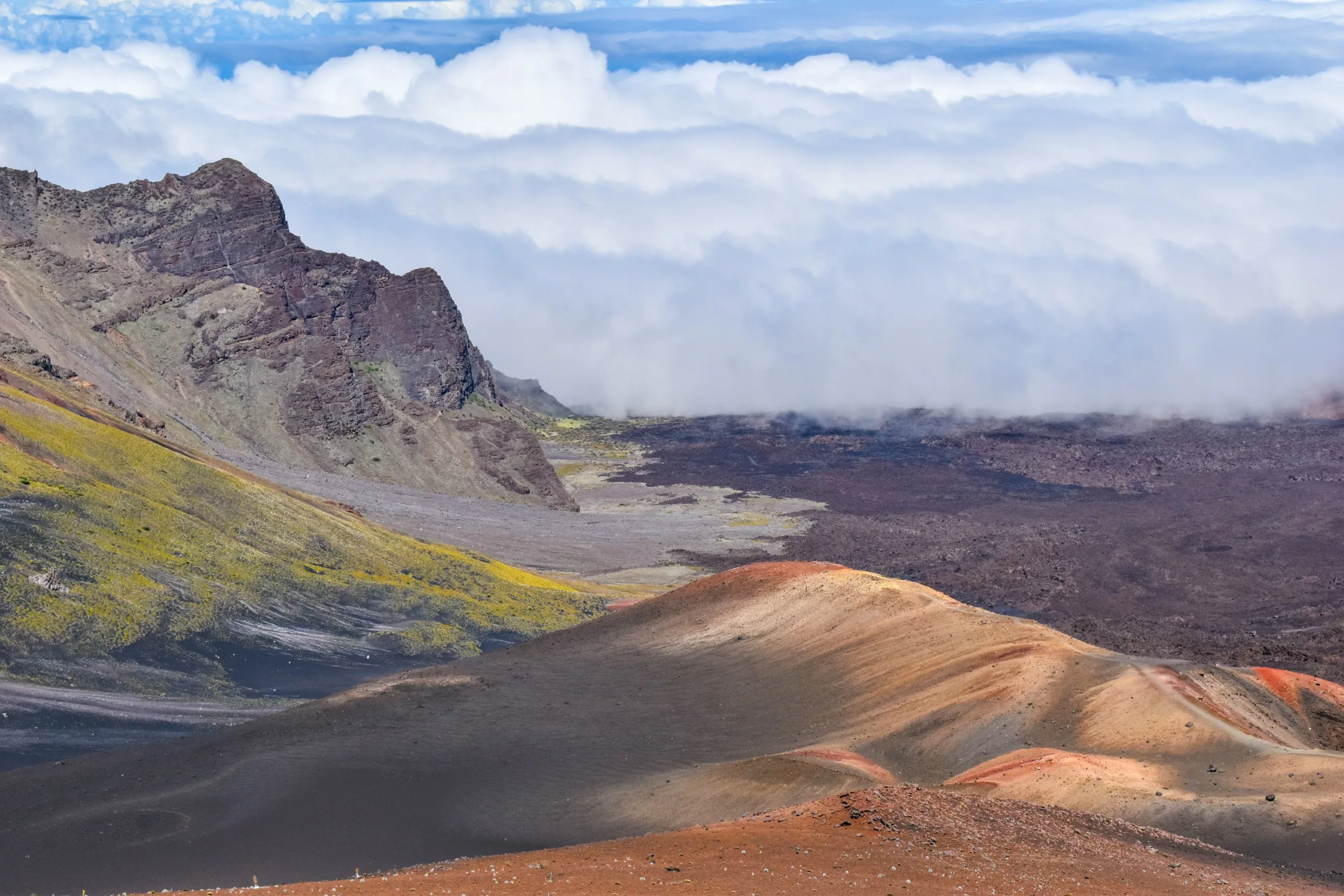 Un paysage volcanique avec des pentes multicolores au premier plan, des falaises rocheuses à gauche et une épaisse couche de nuages couvrant l'horizon