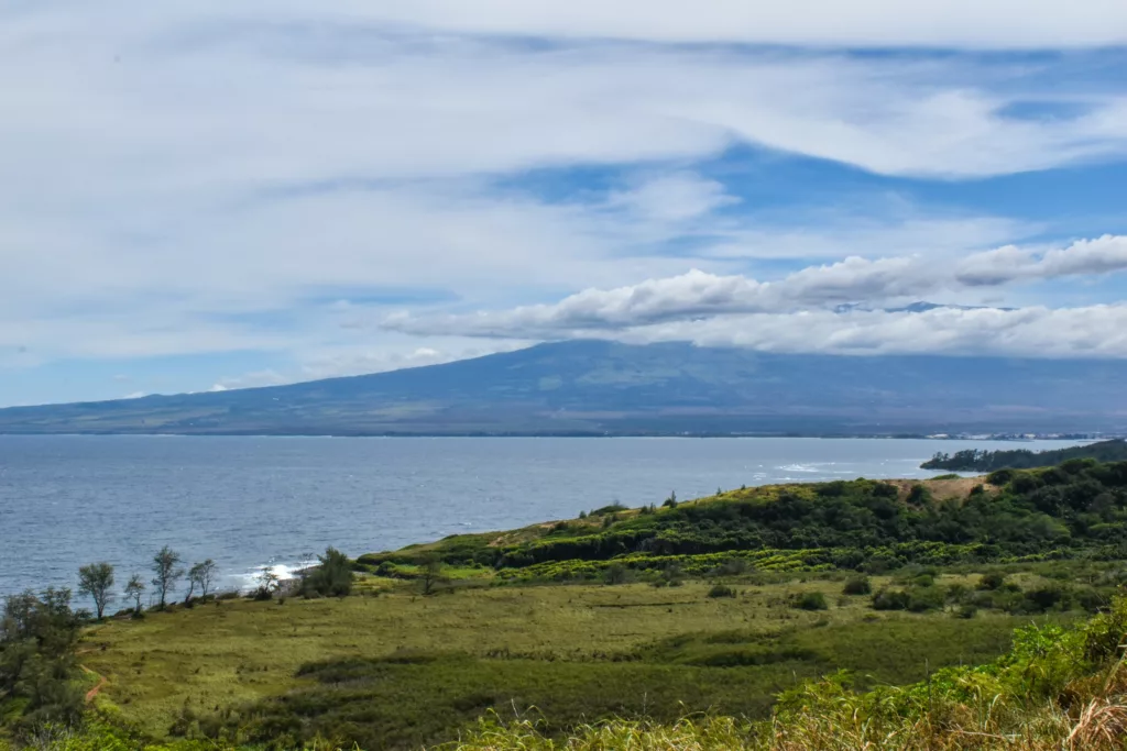 Un paysage côtier avec des collines verdoyantes au premier plan, l'océan au milieu et une grande montagne partiellement couverte de nuages sous un ciel bleu