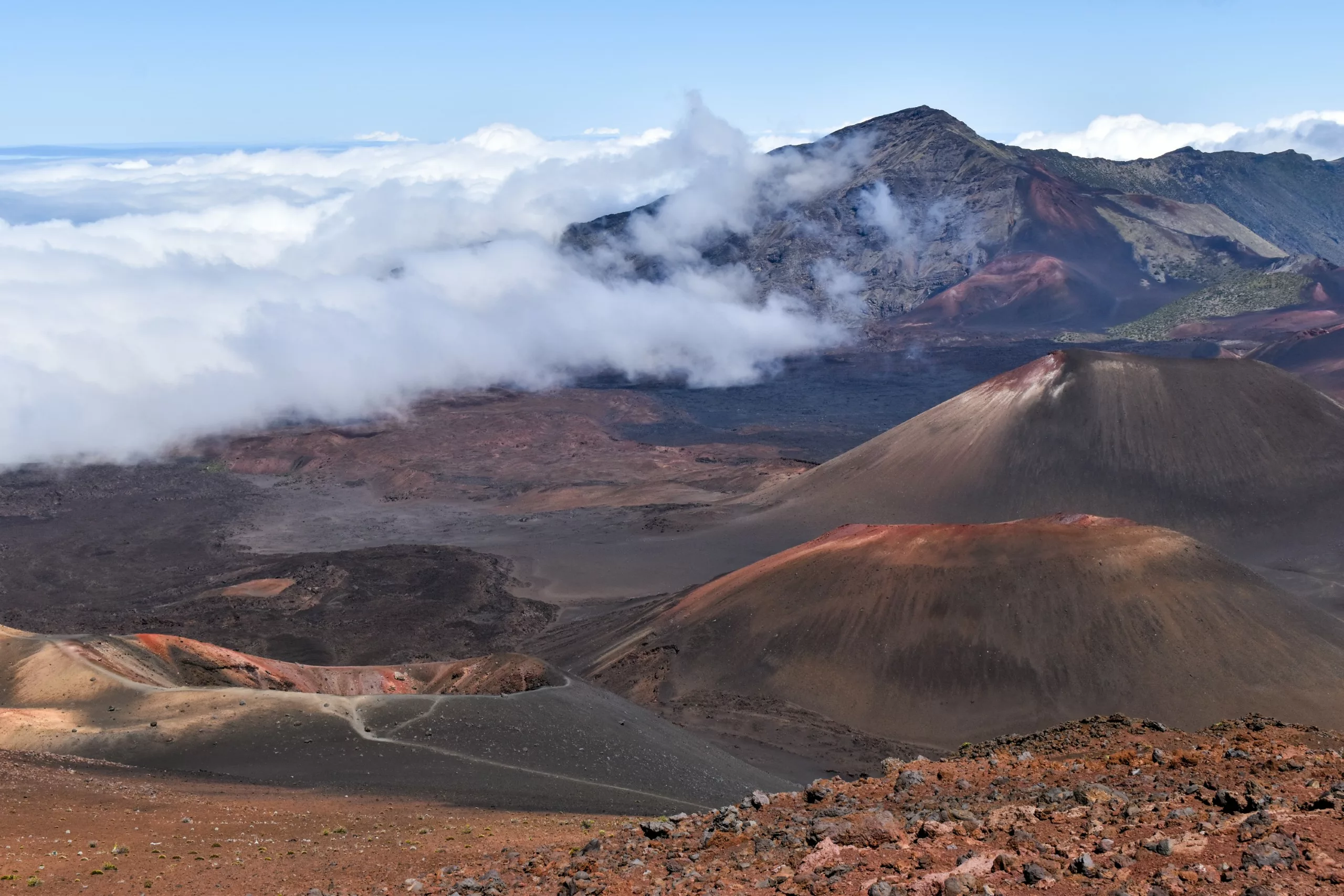 Un paysage volcanique avec plusieurs cônes de scories, un terrain rocheux et une végétation clairsemée sous un ciel bleu. Les nuages passent tandis qu'une montagne s'élève en arrière-plan.