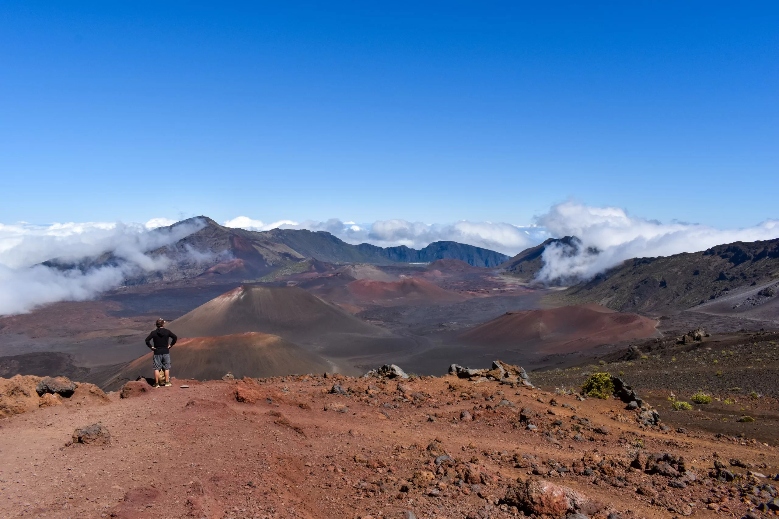 Une personne se tient sur une crête rocheuse rougeâtre surplombant le vaste paysage du cratère volcanique de Haleakala, avec des nuages épars, des montagnes lointaines et un ciel bleu clair en arrière-plan.