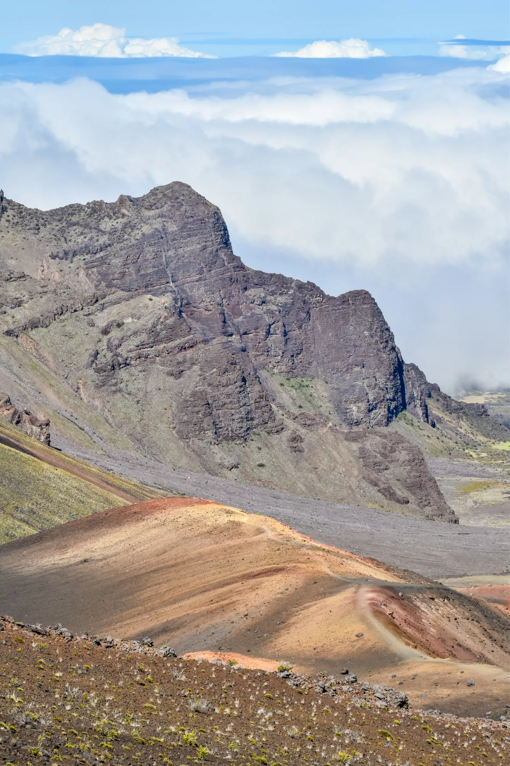 Paysage de montagnes rocheuses avec des pentes brunes et rouges stratifiées au premier plan ; des falaises déchiquetées s'élèvent vers une couche de nuages blancs et de ciel bleu en arrière-plan