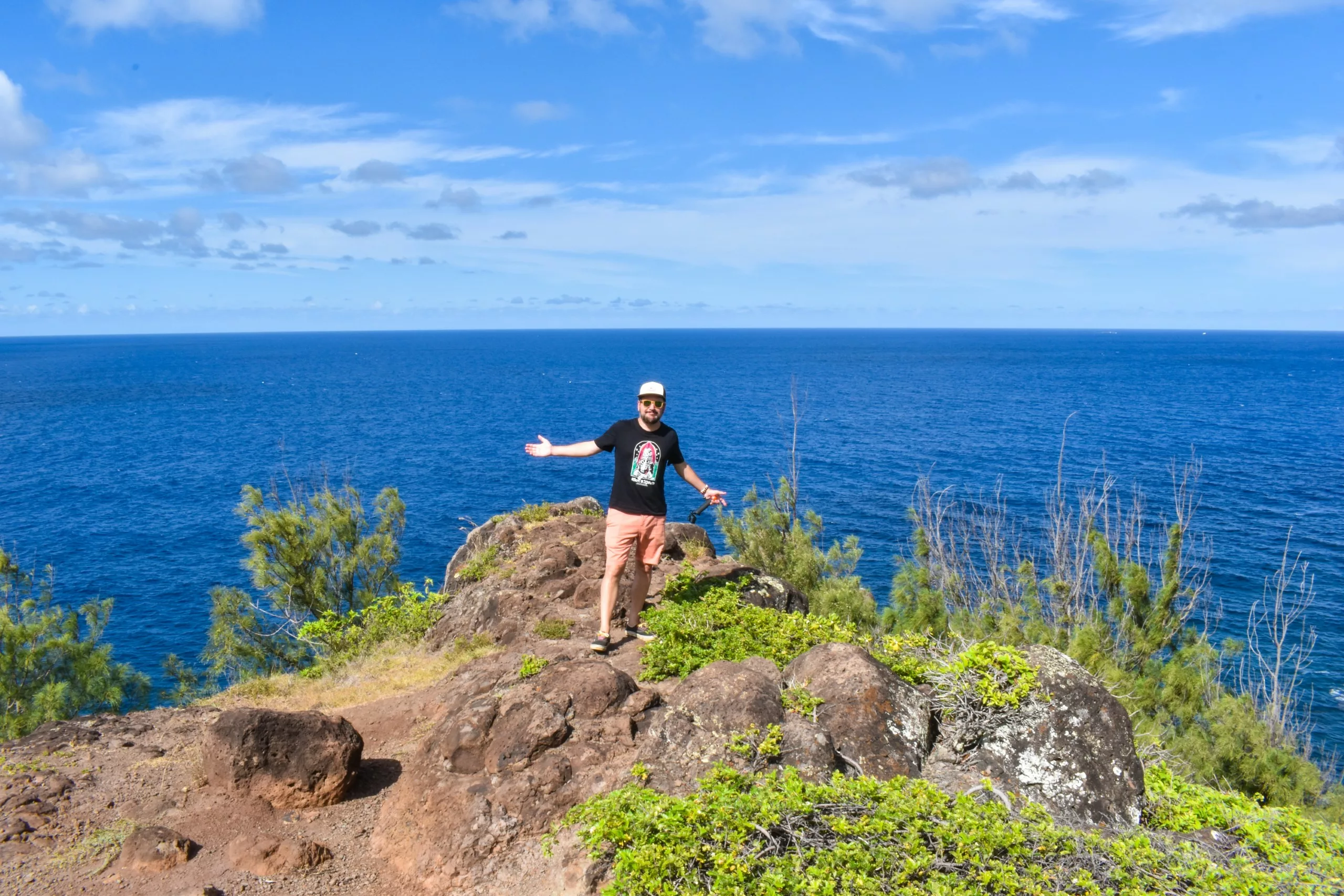Une personne se tient sur une falaise rocheuse, les bras ouverts, surplombant l'eau bleue éclatante de l'océan et un ciel clair - une scène inspirante pour quiconque cherche à faire à Maui parmi des arbustes et des arbres verts luxuriants.