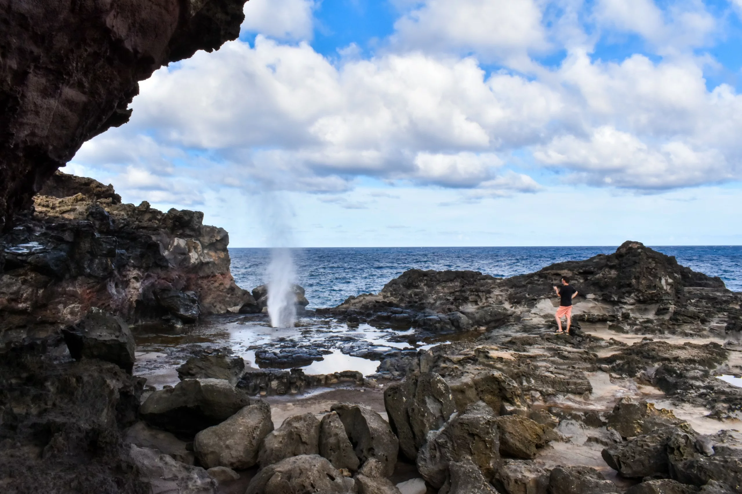 Une personne se tient sur un terrain rocheux près de la côte, observant une trombe d'eau naturelle jaillir