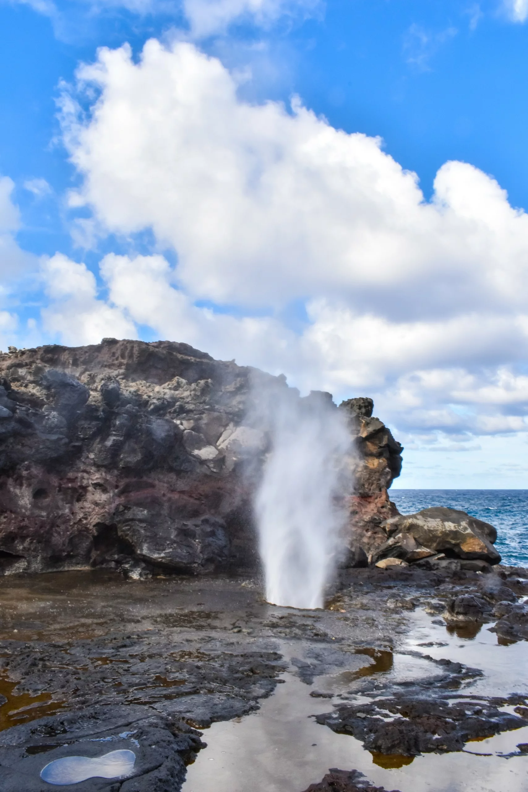 Le trou de soufflage du geyser Nakalele, situé sur un rivage rocheux, projette un jet d'eau dans les airs, avec des nuages dans le ciel et l'océan visible en arrière-plan.