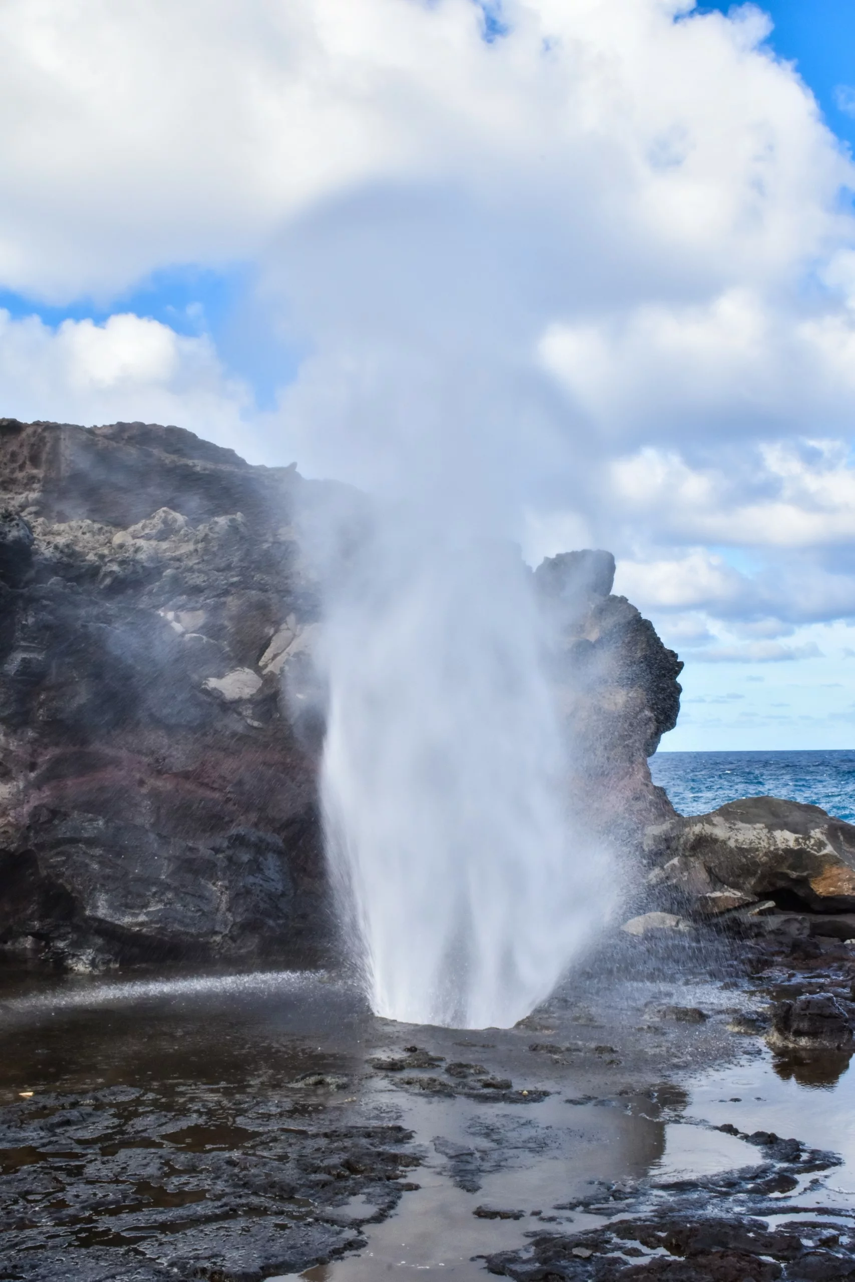 Le geyser de Nakalele projette une haute colonne d'eau et de brume dans les airs à côté de falaises rocheuses sombres, avec l'océan en arrière-plan sous un ciel partiellement nuageux.