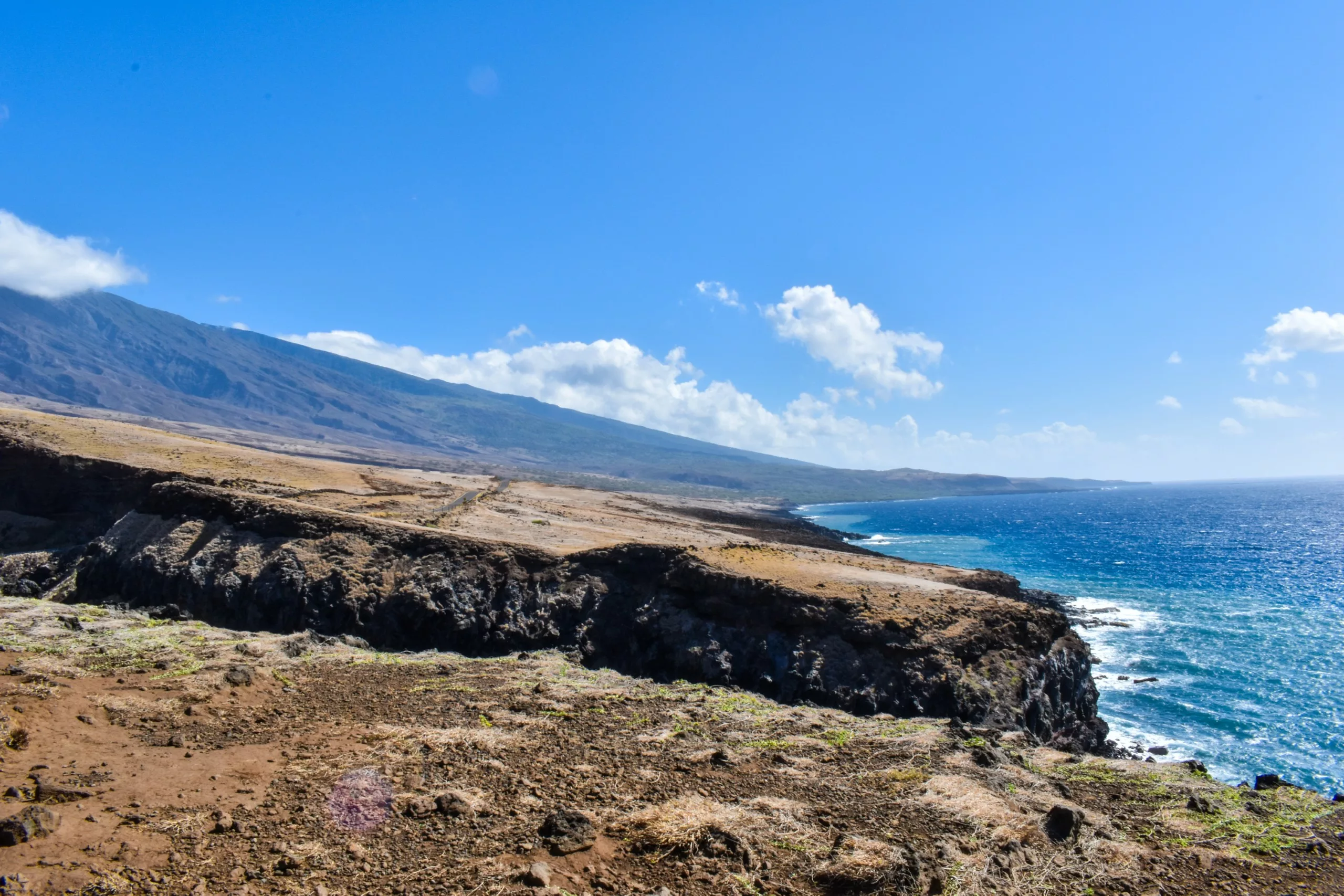 Côte volcanique avec un terrain brun et sec au premier plan. Les falaises rencontrent l'océan à droite et une montagne s'élève à l'arrière-plan sous un ciel bleu clair.