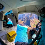 Vue depuis l'intérieur d'un hélicoptère survolant la côte de Kauai avec ses eaux turquoise et ses falaises abruptes ; un visiteur de Kauai portant un casque prend des photos à travers la fenêtre.