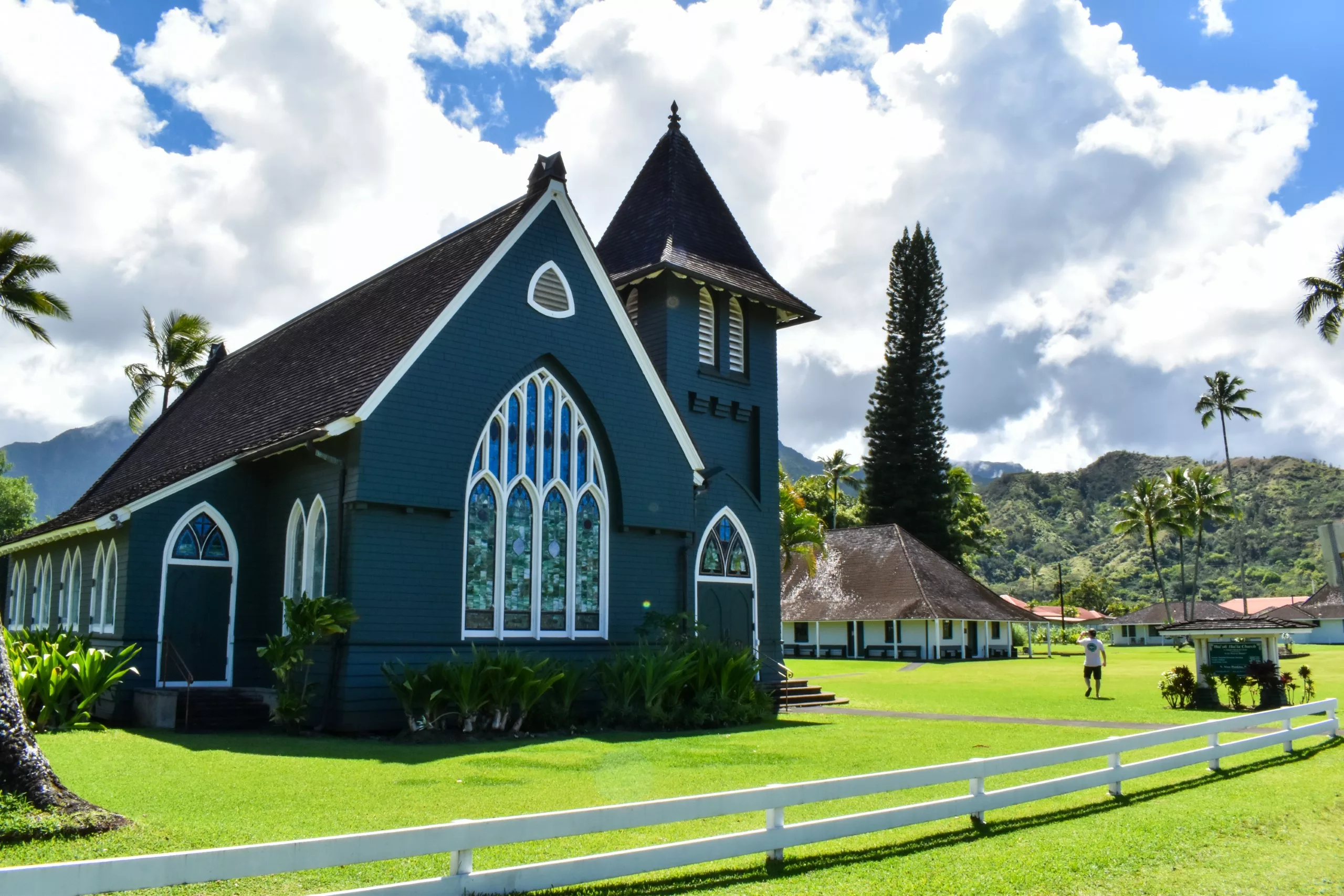 Une église vert foncé, ornée de vitraux et d'un haut clocher, se dresse sur une pelouse verdoyante de Kauai, entourée de palmiers et de montagnes.