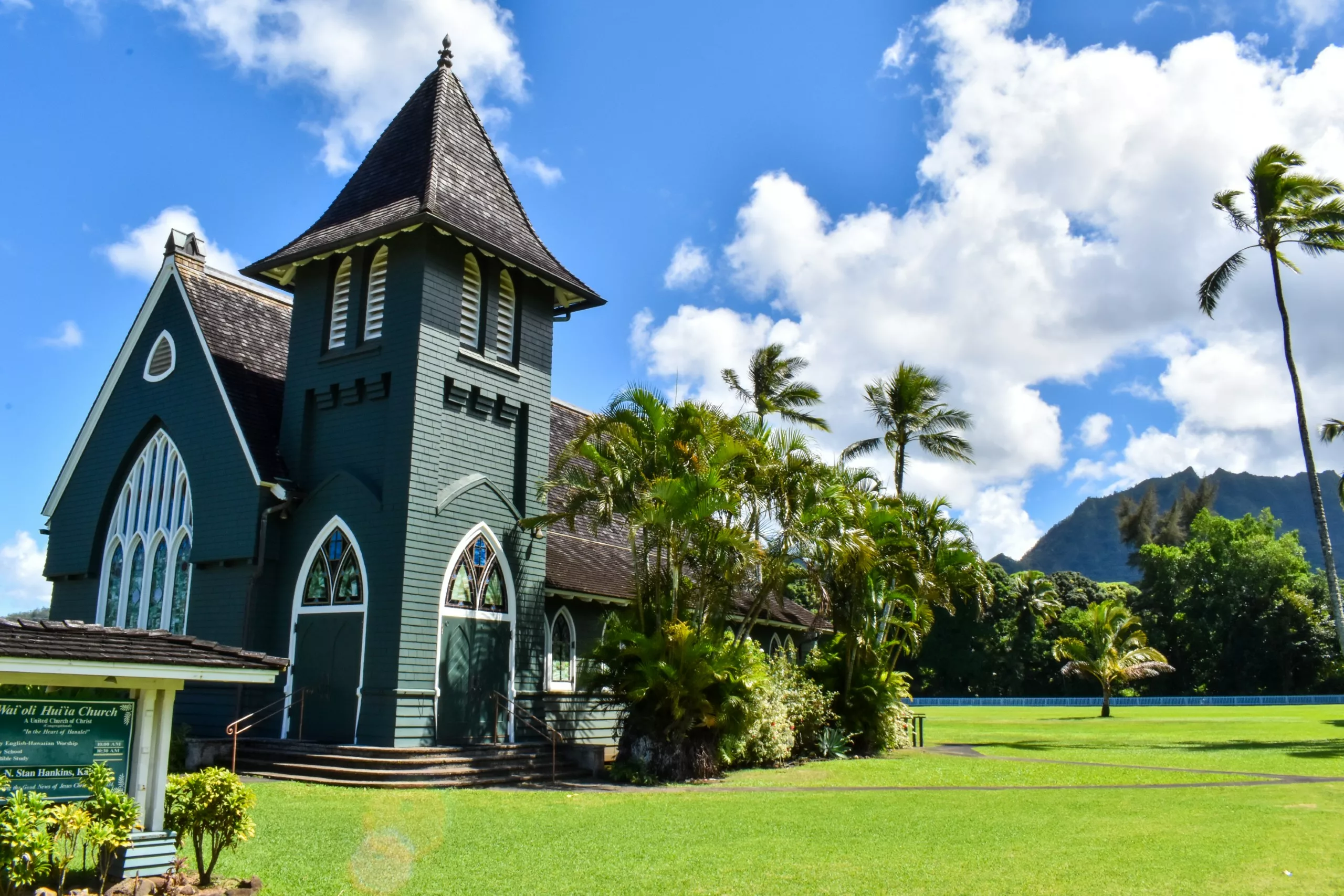 Une église à visiter à Kauai en bois verte avec des vitraux se dresse entourée de palmiers et d'herbe luxuriante sous un ciel partiellement nuageux, avec des montagnes lointaines en arrière-plan.