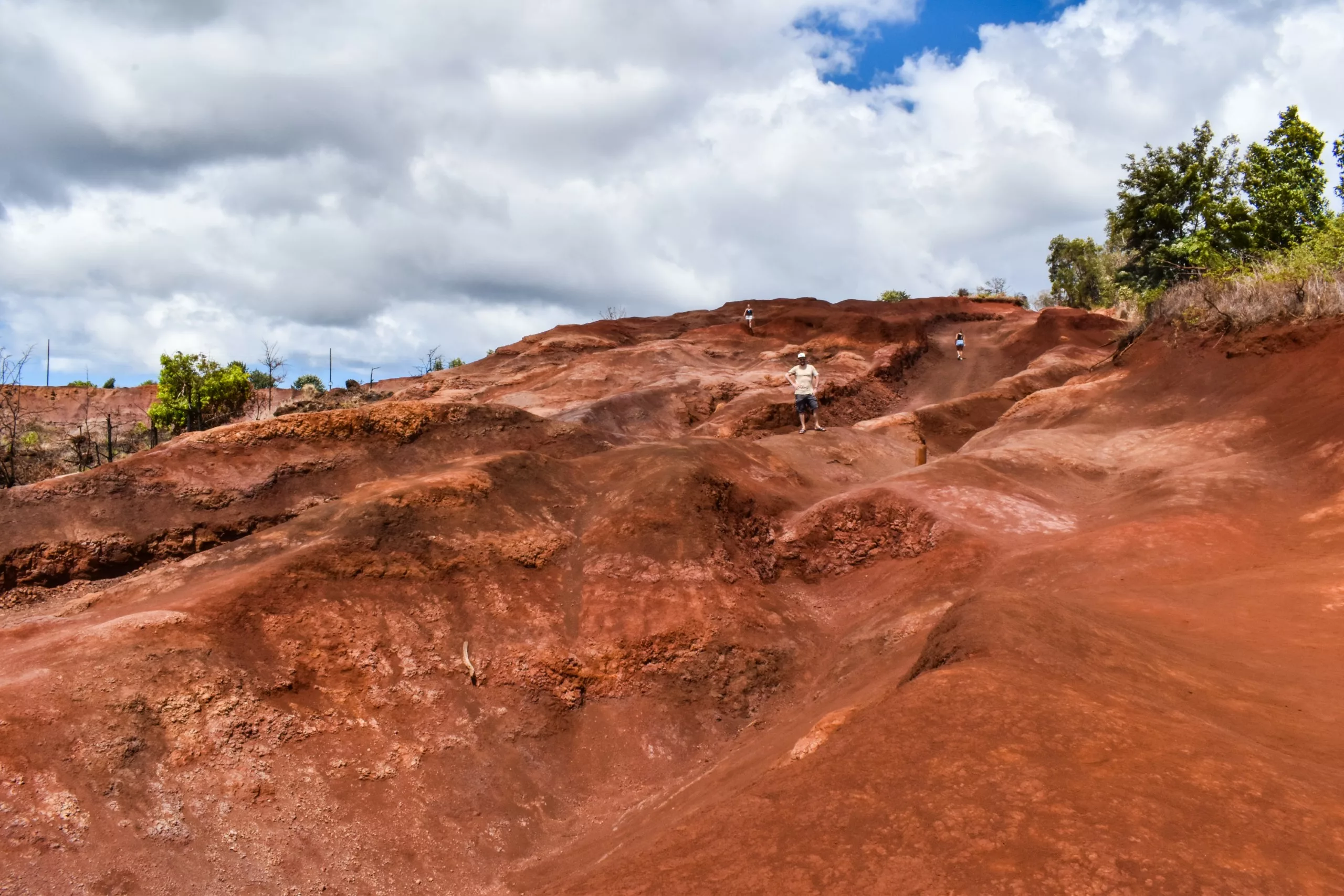 Les gens se tiennent debout et marchent sur des formations rocheuses rougeâtres et inégales sous un ciel nuageux dans ce paysage de Kauai, avec une végétation verte apparaissant le long des bords supérieurs du terrain rocheux.